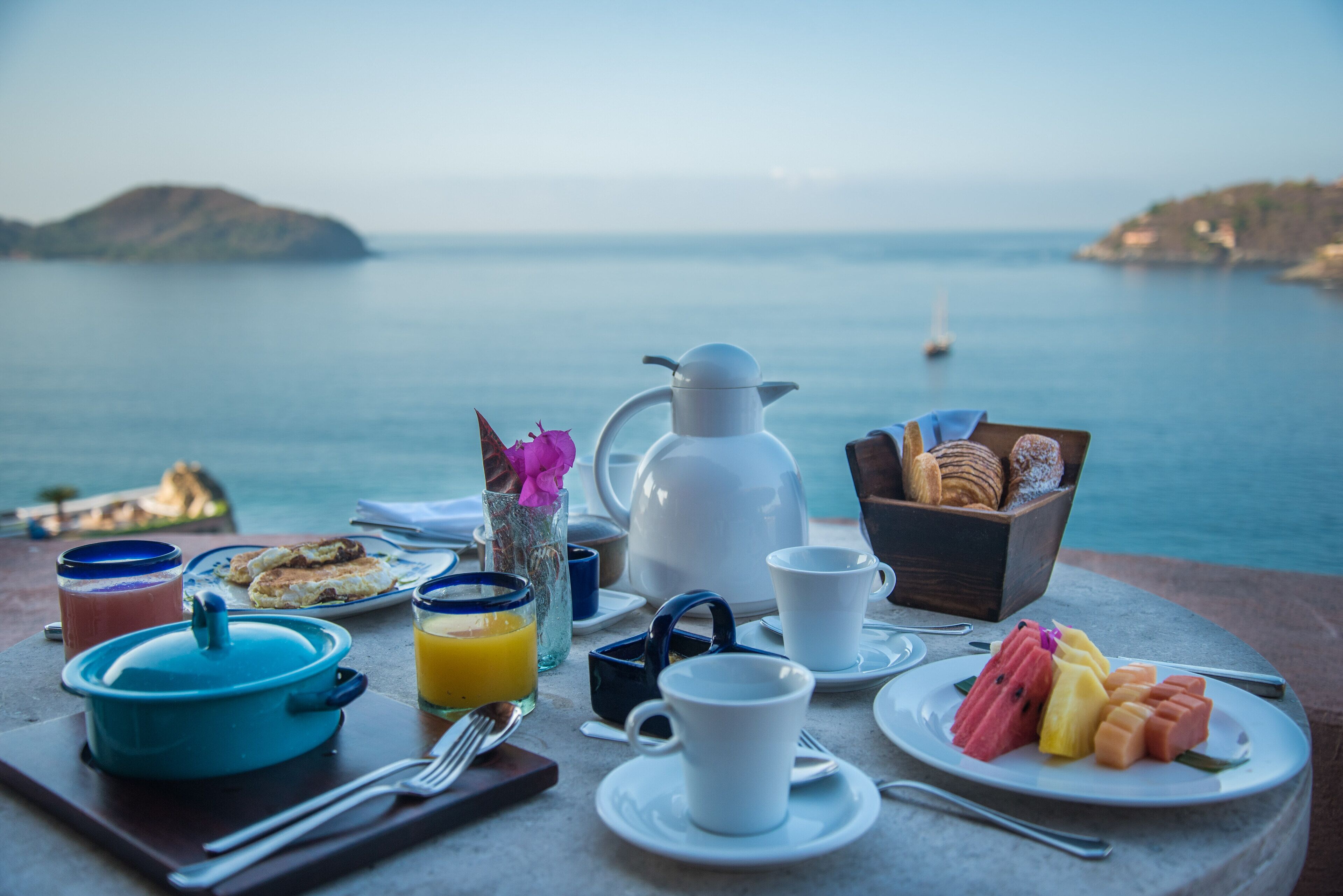 A breakfast table with a view of the sea, including a pitcher, cups, plates, and a box of pastries.