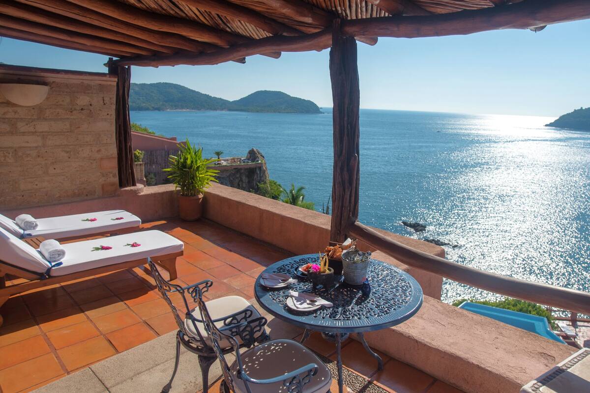 A terrace with two sun loungers, a table with a drink and a flower, and a view of the sea.