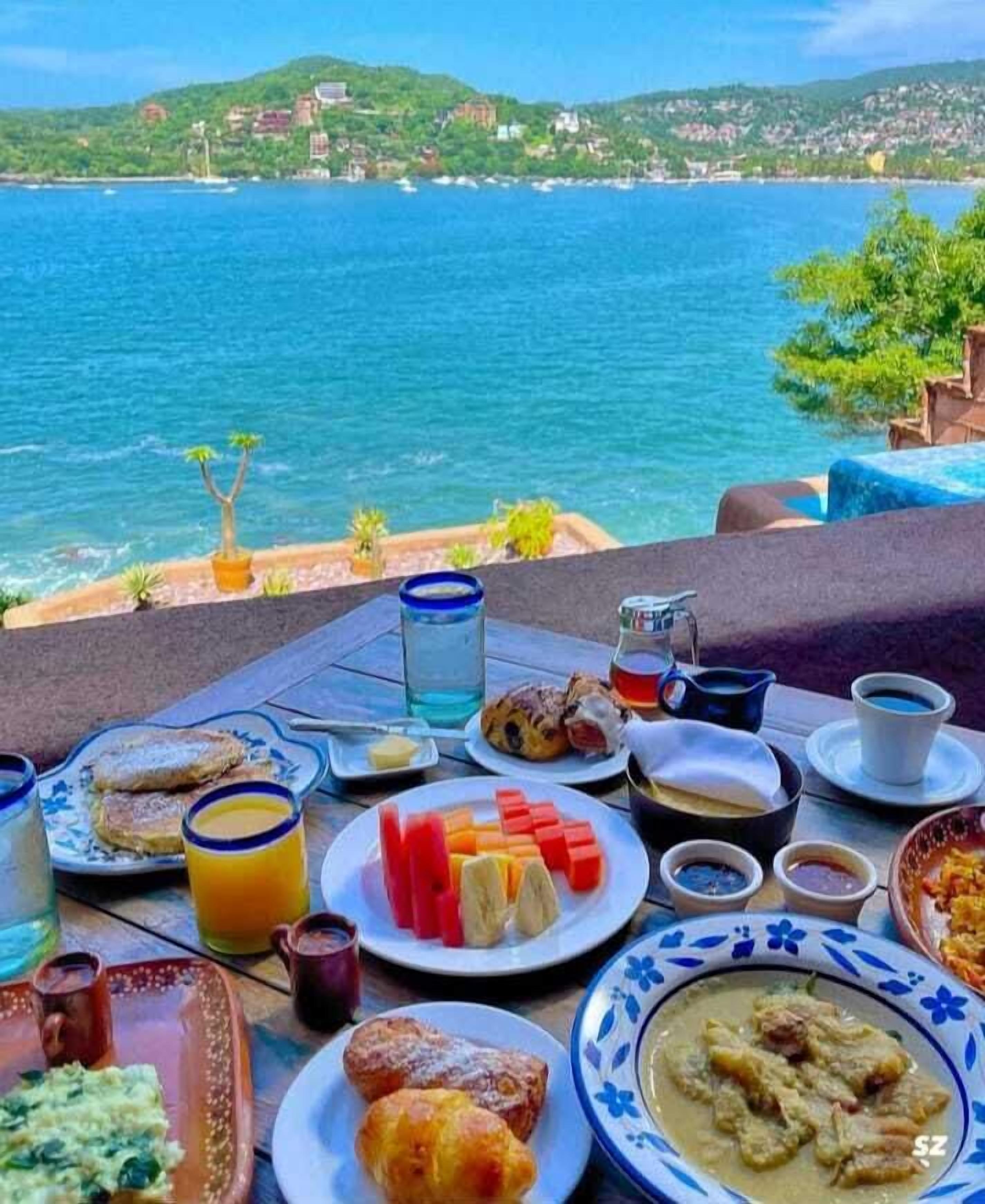 Una mesa servida con alimentos para el desayuno, como tostadas, fruta, pasteles y un bol de sopa, con vistas a un paisaje costero.