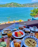 Una mesa servida con alimentos para el desayuno, como tostadas, fruta, pasteles y un bol de sopa, con vistas a un paisaje costero.