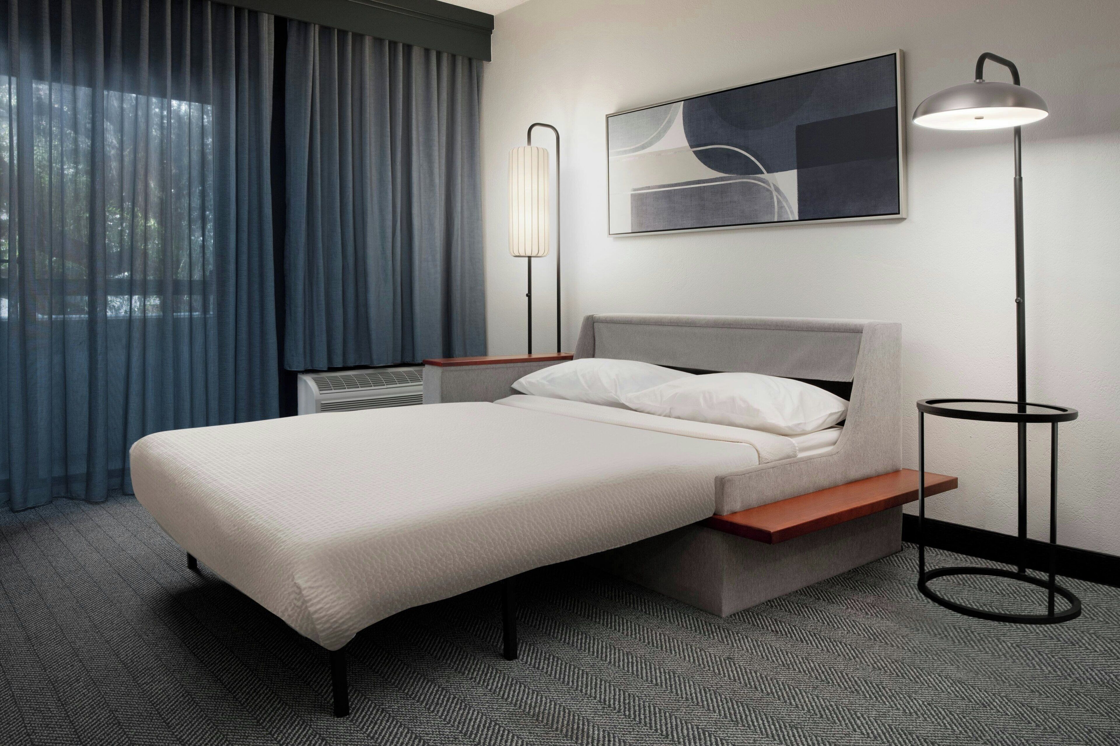 A modern hotel room with a bed, a bedside table, a lamp, and a window with curtains.
