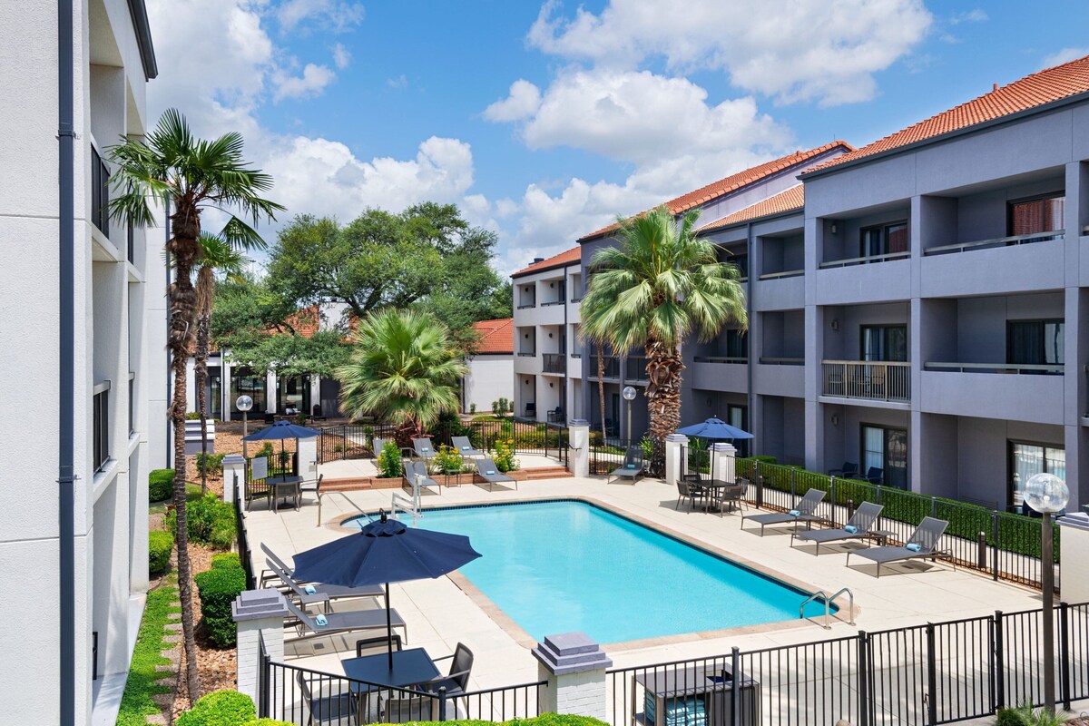 A swimming pool surrounded by lounge chairs and umbrellas, with palm trees and a multi-story building in the background.