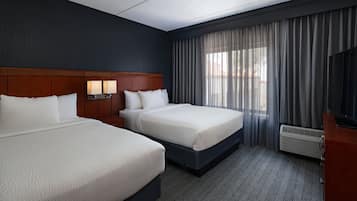 A hotel room with two beds, a wooden headboard, a nightstand with a lamp, a TV, and a large window with curtains.