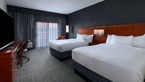 A hotel room with two beds, a desk with a computer, a TV, and a large window with curtains.