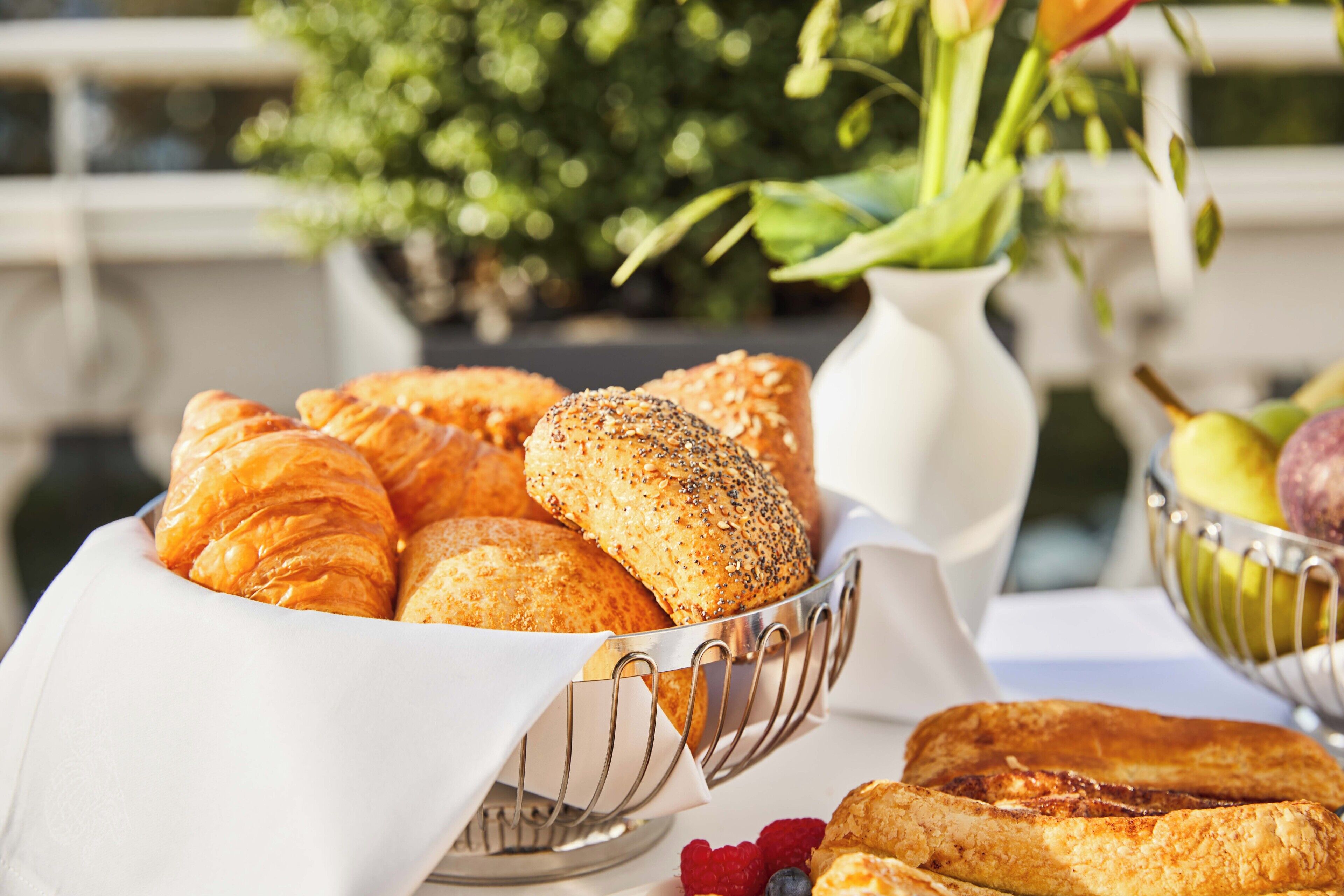 A table with a variety of baked goods including croissants and bread rolls, accompanied by fresh fruits and a vase with flowers.