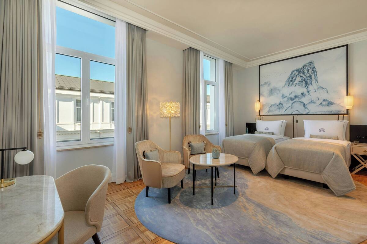 Habitación de hotel con un ventanal grande, dos sillones, una mesa redonda, una cama con dos almohadas y un cuadro en la pared.