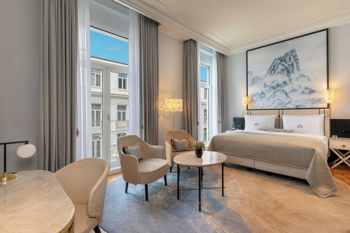 Habitación de hotel con cama, dos sillas, una mesita, una lámpara y un cuadro grande en la pared.