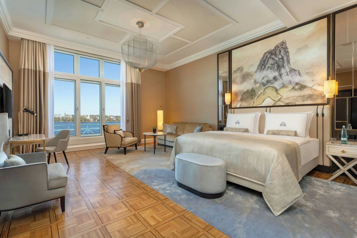 Amplia habitación de hotel con una cama grande, una zona de estar con una mesa y sillas, un ventanal con vistas y un cuadro en la pared.
