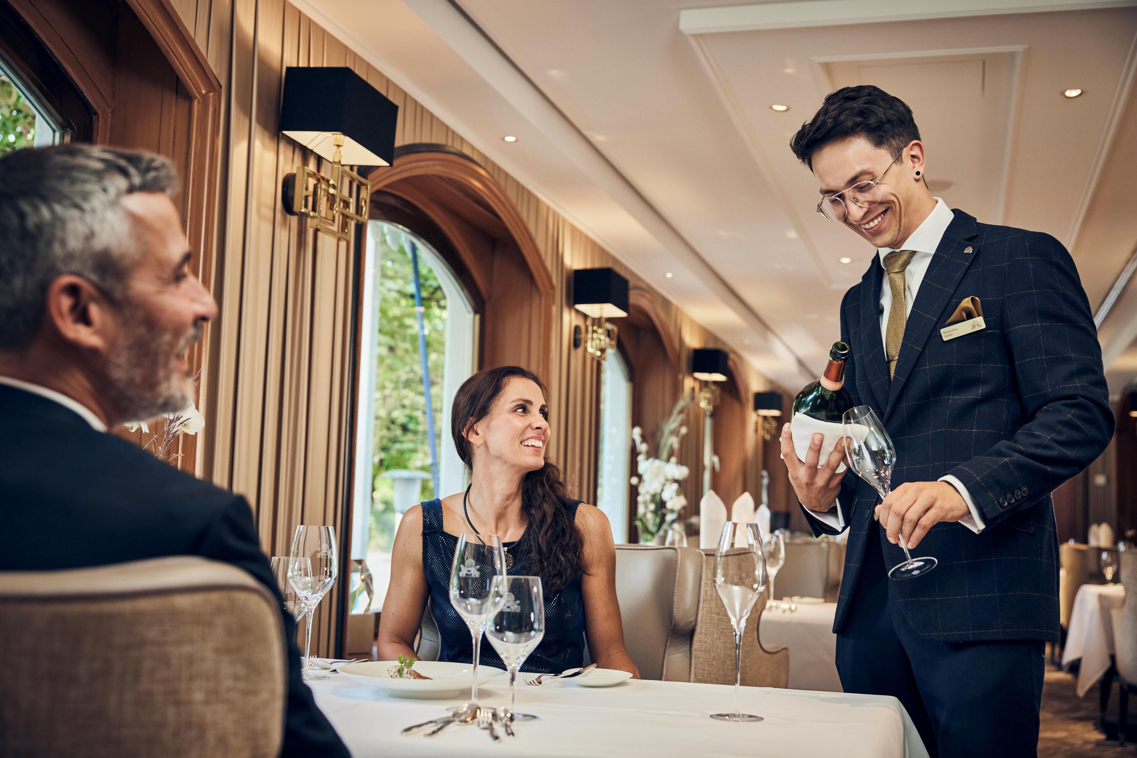Een eetgedeelte met een tafel voor drie personen, een ober met een fles wijn, en een raam met uitzicht op groen.