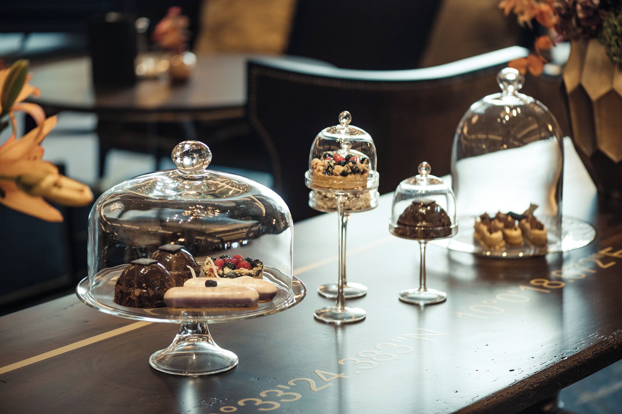 Une table sur laquelle sont disposés divers desserts sous des cloches de verre, une table en bois portant le numéro « 3334 » gravé, et un arrière-plan flou avec une chaise et une plante.