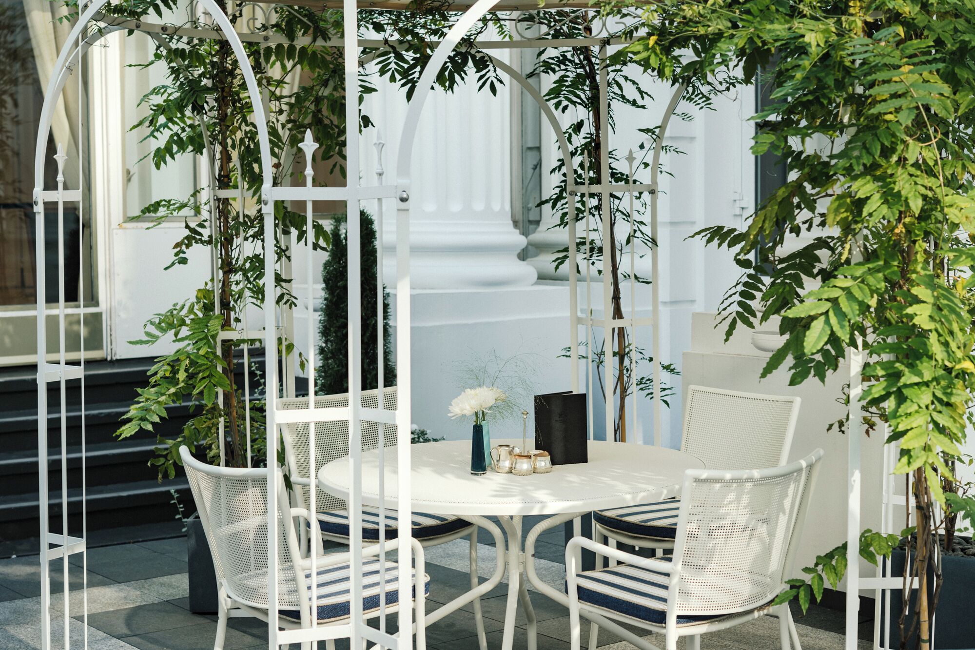 Un espace extérieur aménagé avec des sièges blancs, comprenant une table ronde et des chaises, le tout entouré de rampes de protection en métal blanc et de verdure.