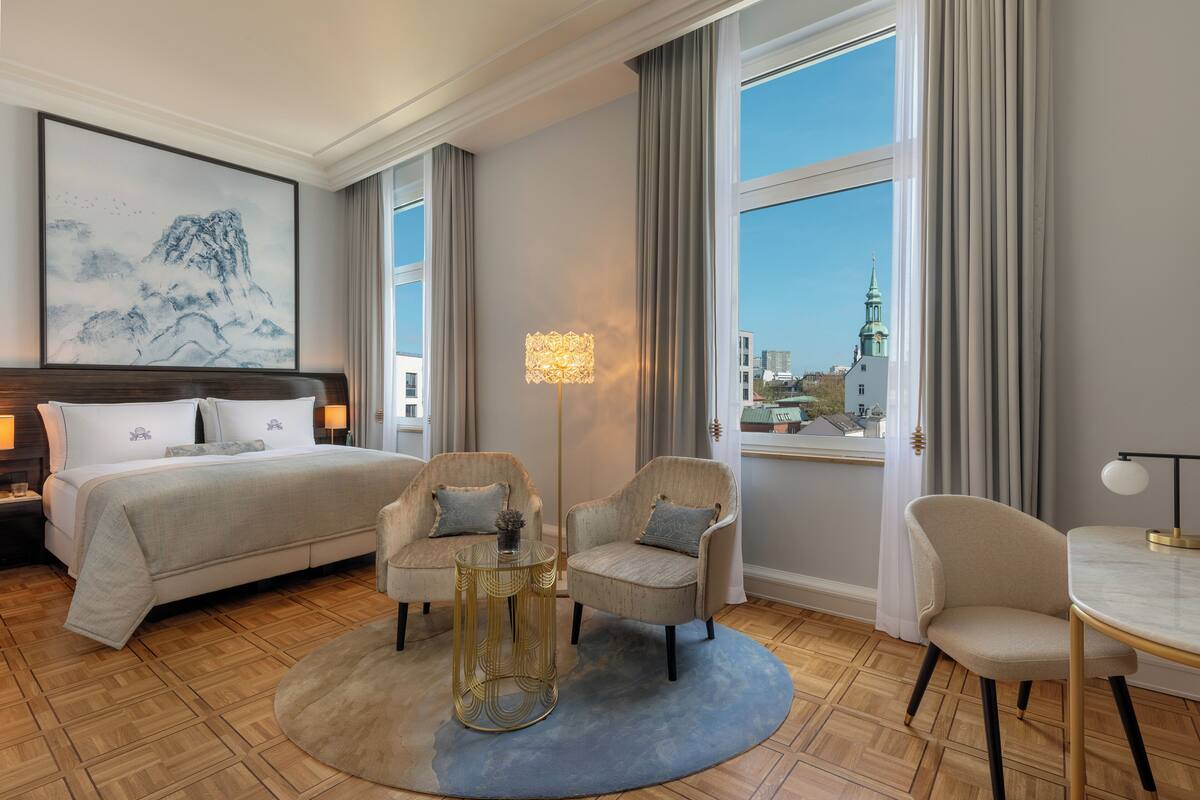 Habitación de hotel con cama, dos sillones, una mesa de centro redonda, una lámpara y un amplio ventanal con vista a la ciudad.