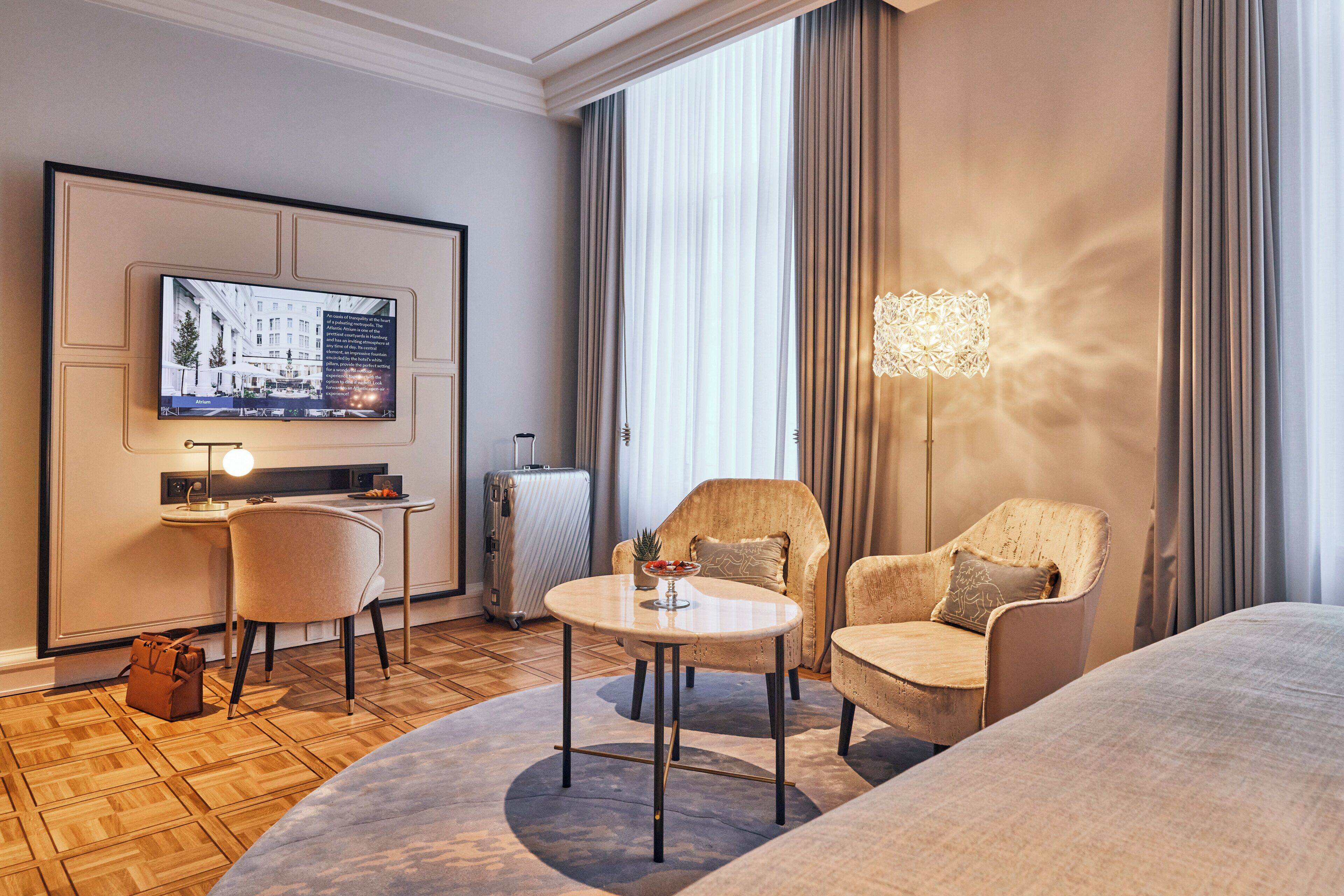 Een hotelkamer met een flatscreen televisie, een ronde tafel, twee fauteuils, een bank, een koffer en een lamp.