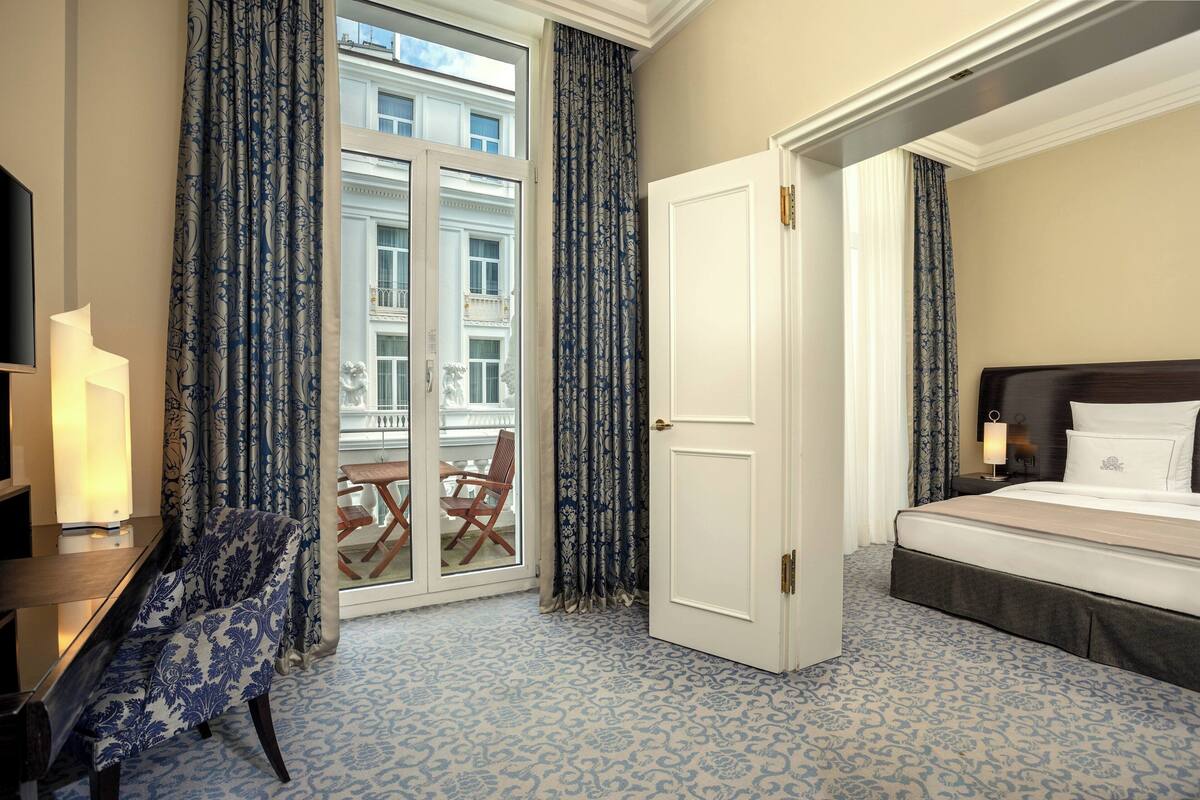 Habitación de hotel con cama, una silla, escritorio y balcón con vista a edificios.