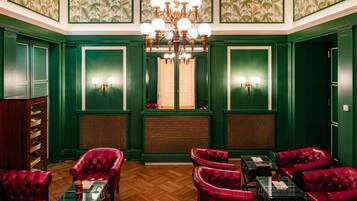 Una habitación con paredes verdes, piso de madera y sillones de cuero rojo. Se observa un candelabro, un ventanal con vistas y una puerta.