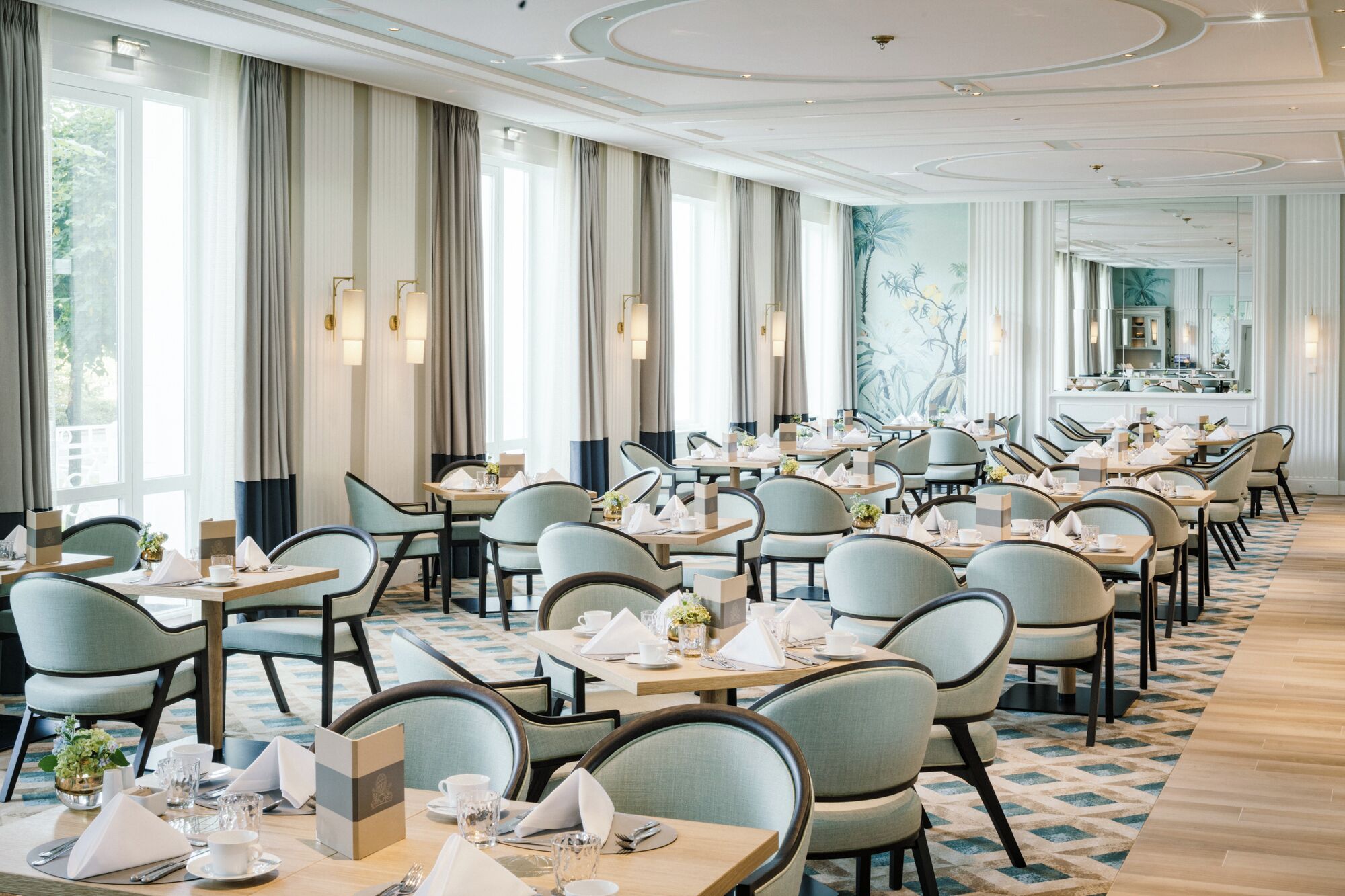 A spacious dining area with multiple tables and chairs, large windows with curtains, and a patterned carpet.