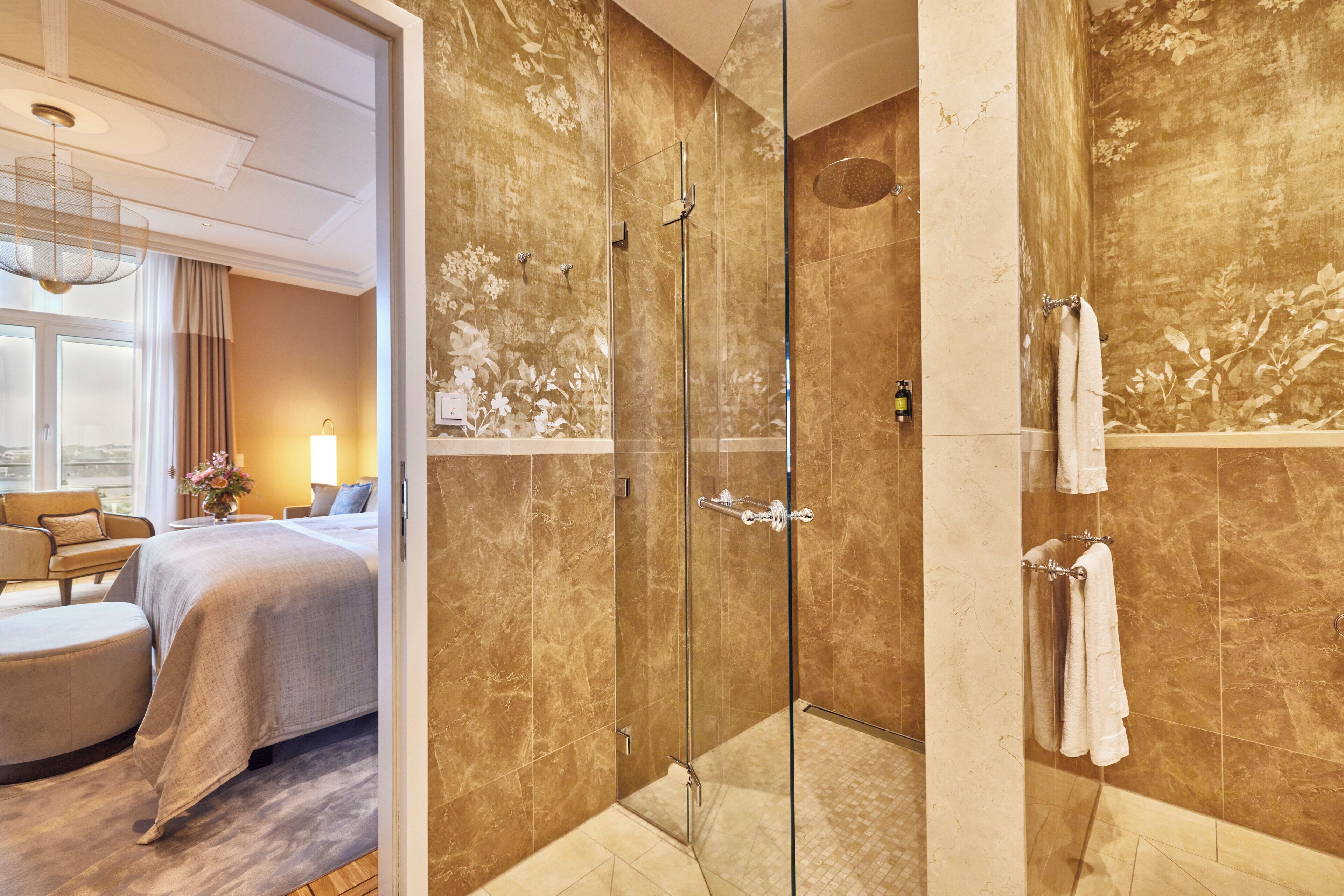A hotel room with a glass-enclosed shower, a bed with a beige bedspread, a sitting area with a chair and ottoman, and a view of the outdoors.