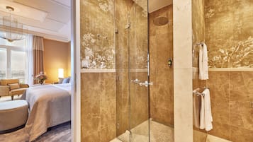 A hotel room with a glass-enclosed shower, a bed with a beige bedspread, a sitting area with a chair and ottoman, and a view of the outdoors.