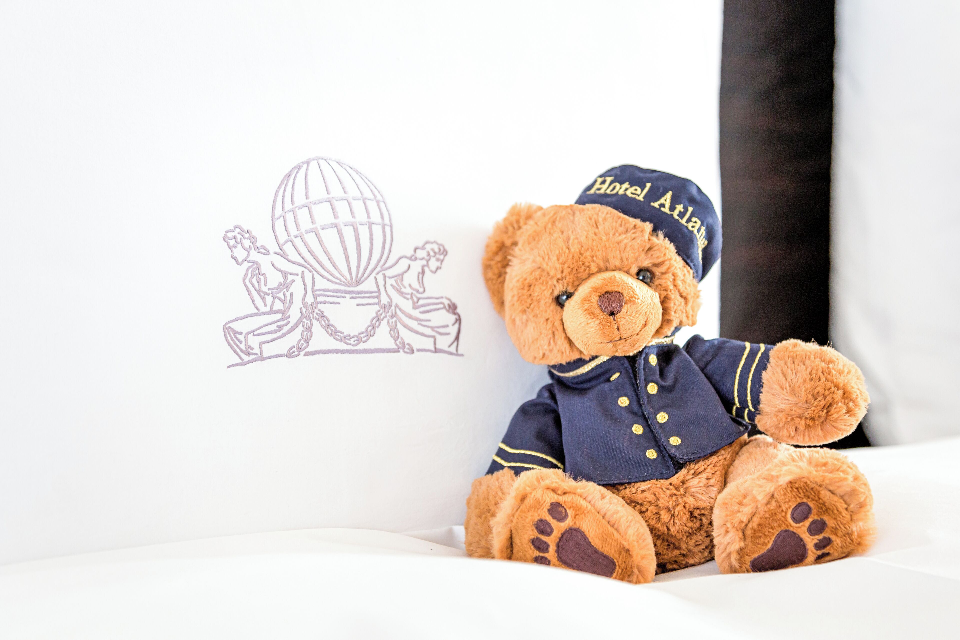 Un ours en peluche, vêtu d’un costume de marin, est assis sur une surface blanche, à côté d’un mur orné d’un dessin représentant un globe terrestre et une personne.