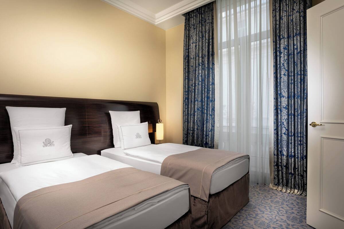 Habitación de hotel con dos camas, cabecero de madera, ropa de cama blanca y cortina estampada.