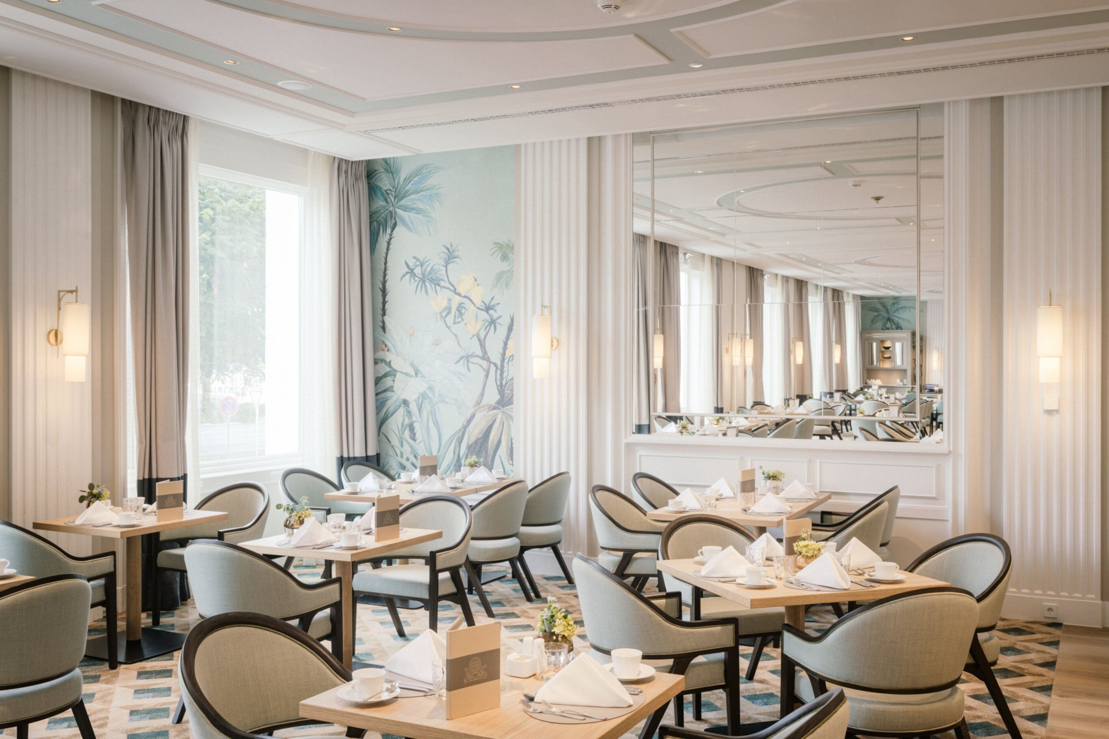 Une salle à manger d’hôtel avec des tables dressées pour le service, de grandes fenêtres et une fresque murale.