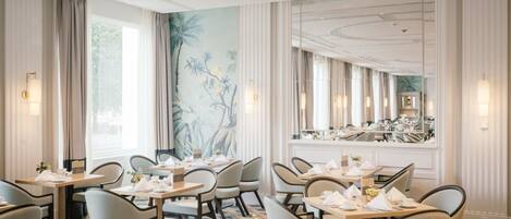 Une salle à manger d’hôtel avec des tables dressées pour le service, de grandes fenêtres et une fresque murale.