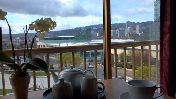 Une pièce avec vue sur un paysage urbain, comprenant un bâtiment moderne et un pont. On y trouve une table avec une théière, des tasses et une plante en pot.