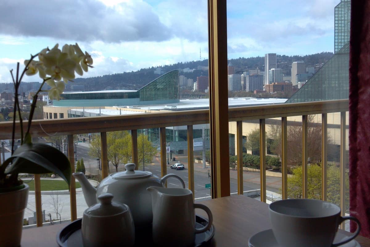 A room with a view of a cityscape, including a modern building and a bridge. There is a table with a teapot, cups, and a potted plant.