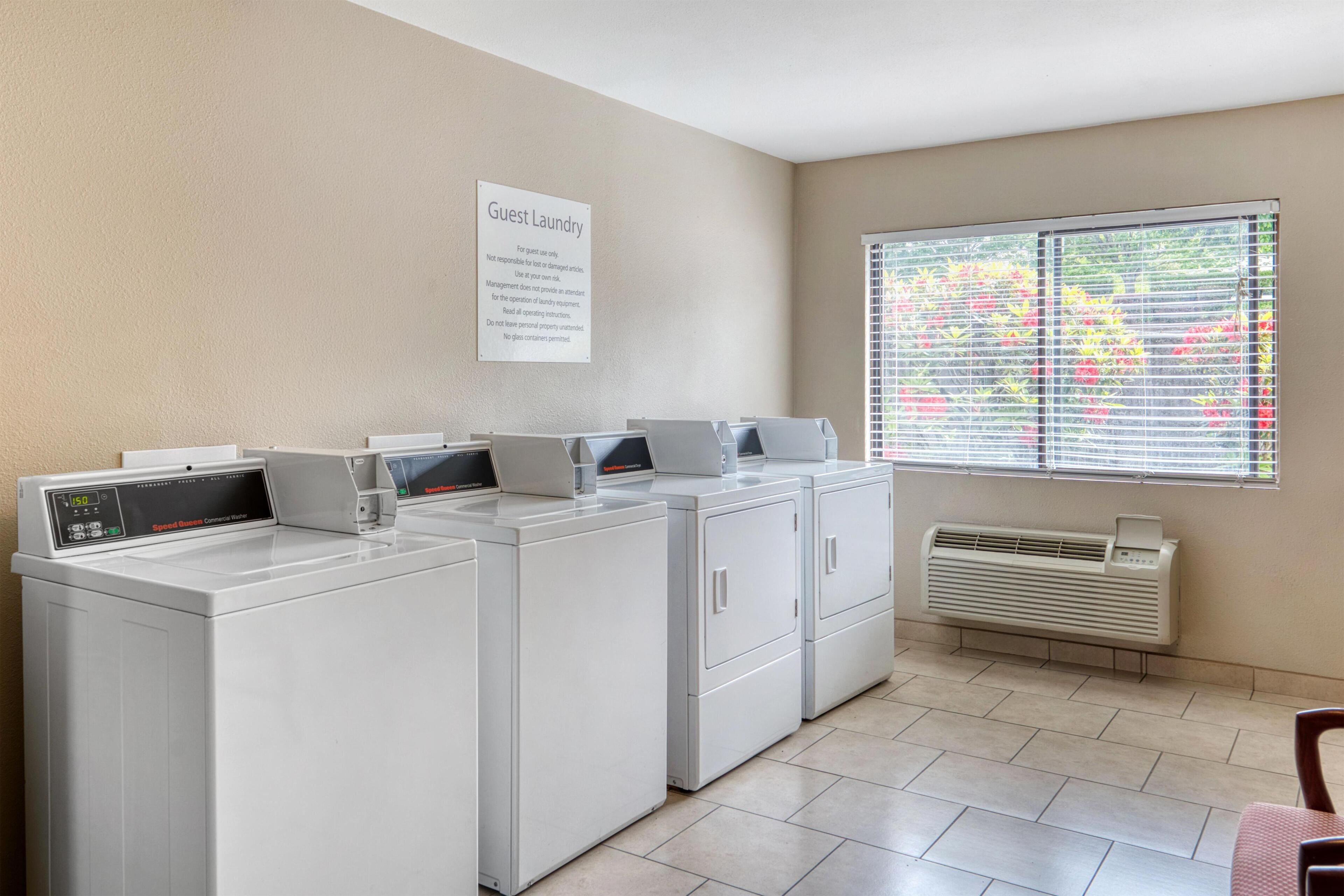 A laundry area with four washing machines, a sign indicating guest laundry, a window with blinds, and a wall-mounted air conditioner.