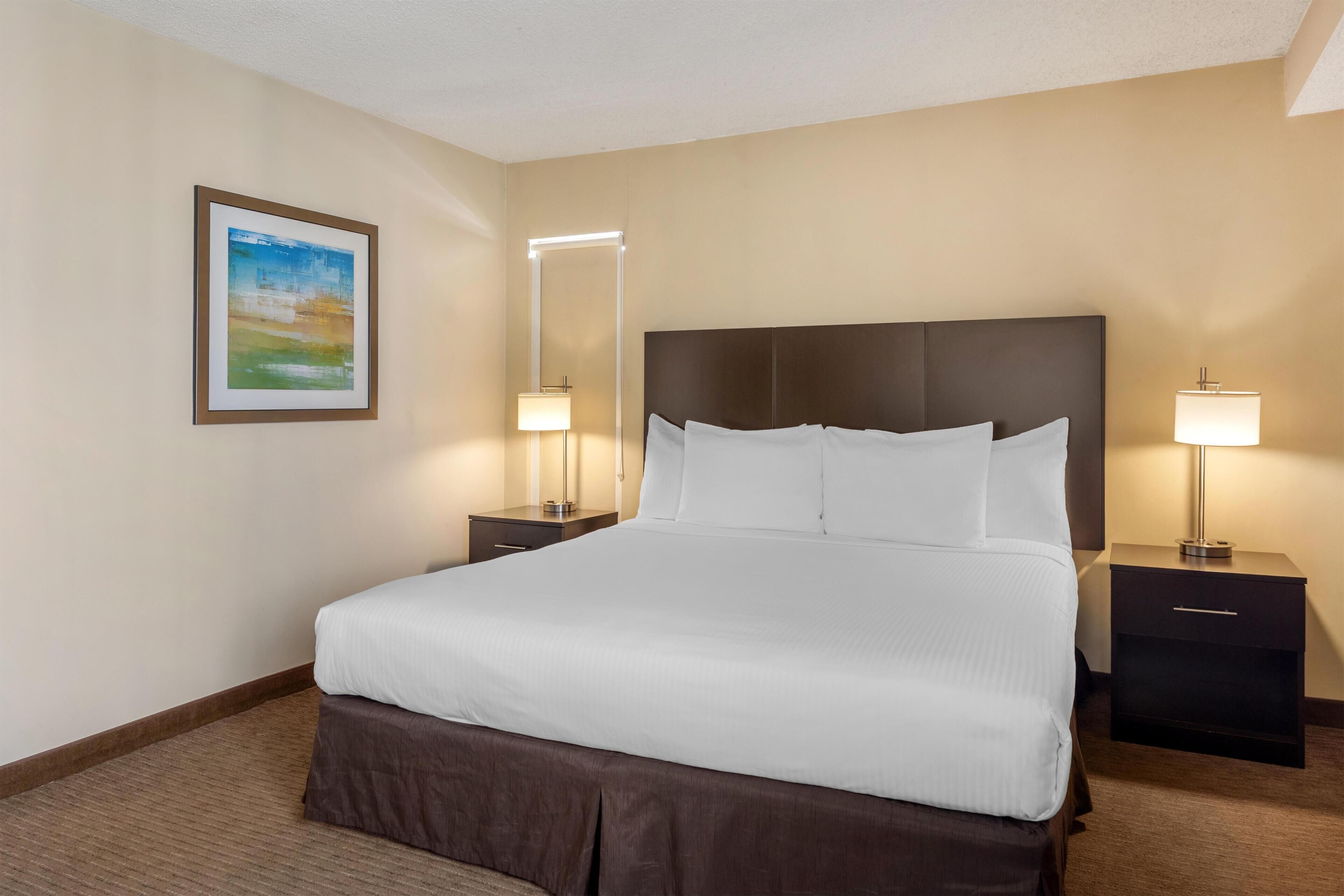 A hotel room with a large bed, two bedside tables, a nightstand, a wall-mounted lamp, and a framed picture.
