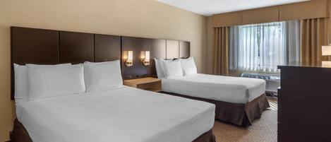 A hotel room with two beds, a desk, a chair, a window with curtains, and a wall-mounted lamp.