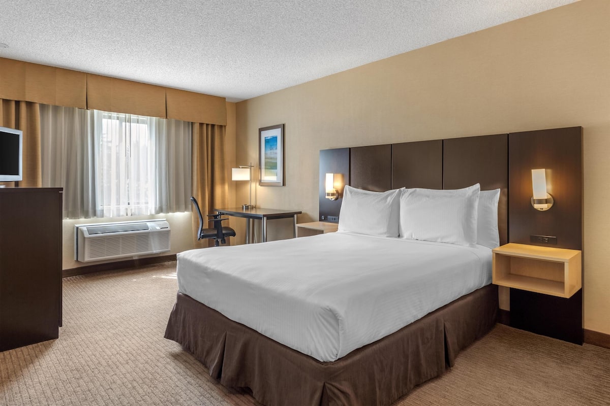 Ein Hotelzimmer mit einem großen Bett, einem Schreibtisch, einem Sessel, einem Fernseher und einem Fenster mit Vorhängen.