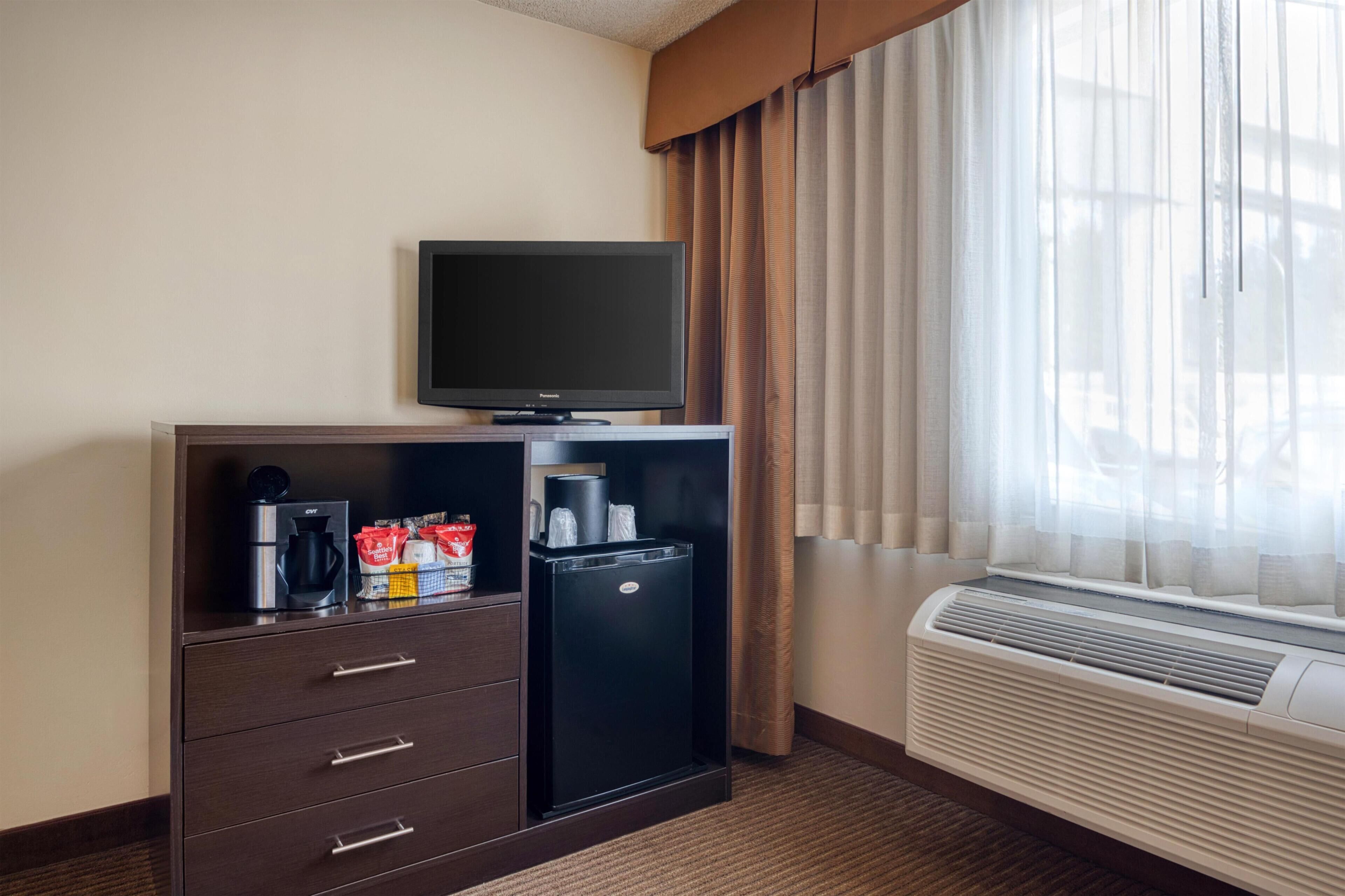 A hotel room with a flat-screen TV, a minibar, a coffee maker, and a window with curtains.
