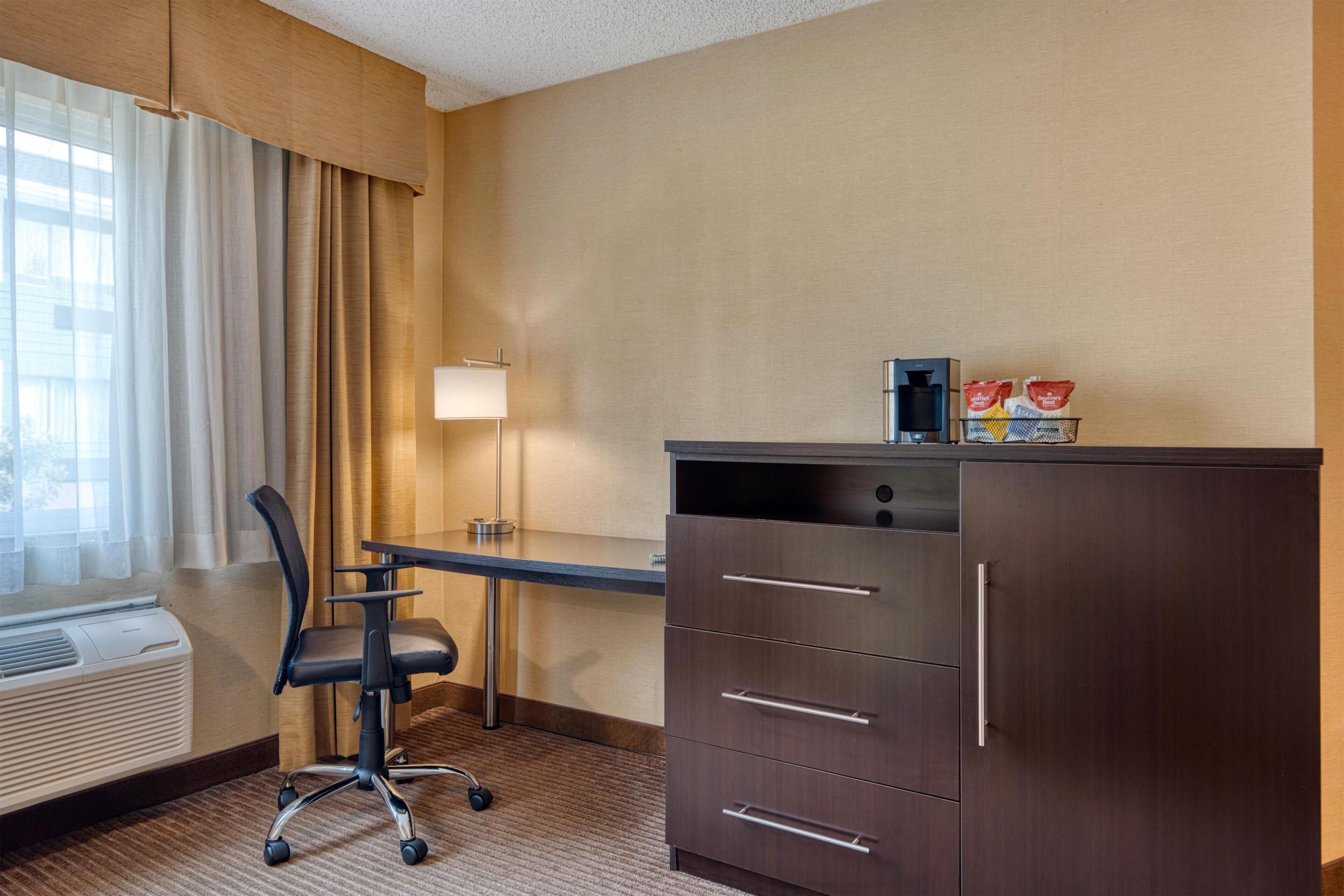 Habitación de hotel con escritorio, silla, armario, lámpara y ventana con cortinas.
