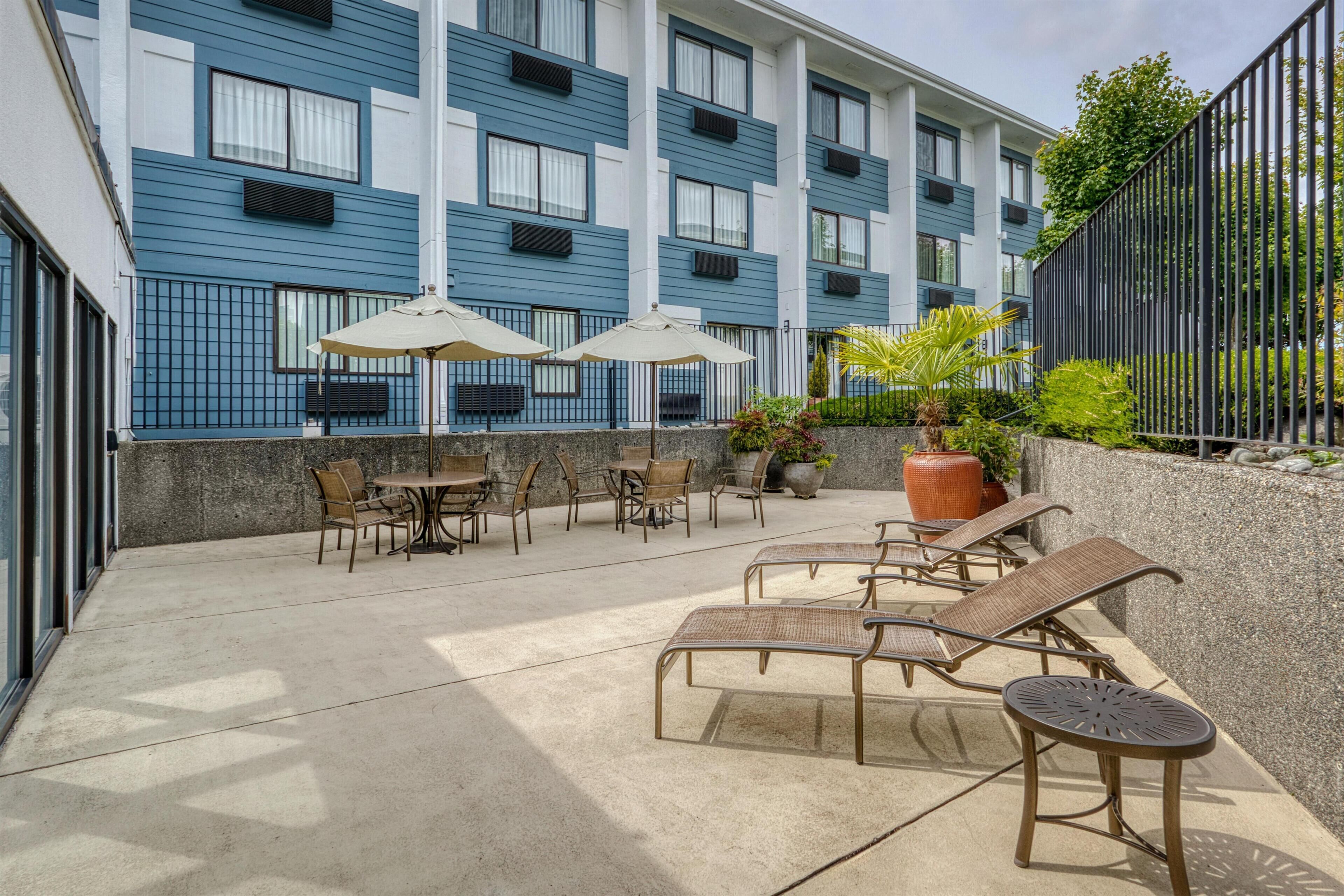 An outdoor patio with lounge chairs and umbrellas, a concrete floor, and a building with blue and white exterior walls.