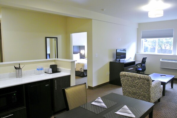 A hotel room with a kitchenette, a dining area with a table and chairs, a TV, and a window with blinds.