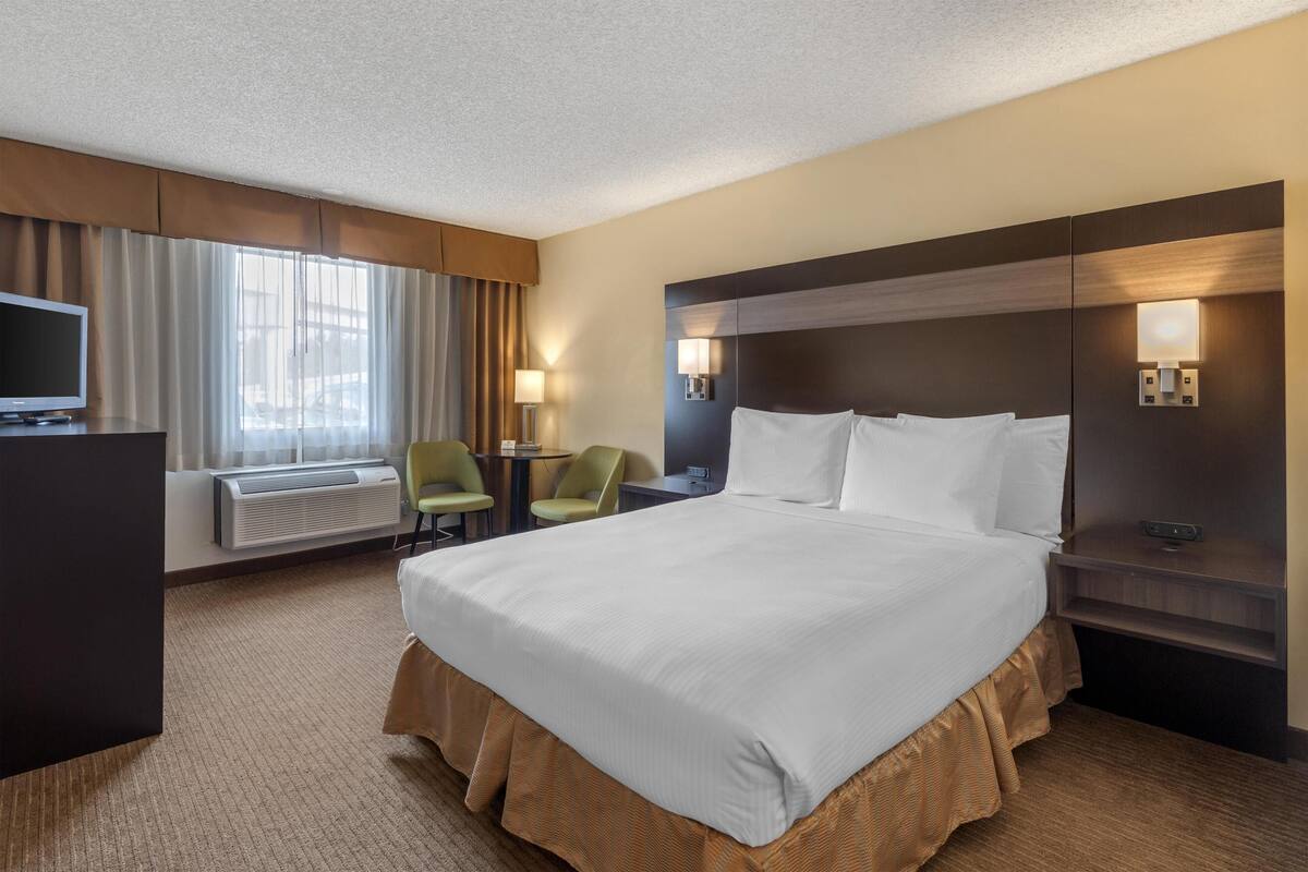 Ein Hotelzimmer mit einem großen Bett, einem Schreibtisch, einem Flachbildfernseher, einem Sessel, einer Lampe und einem Fenster mit Vorhängen.
