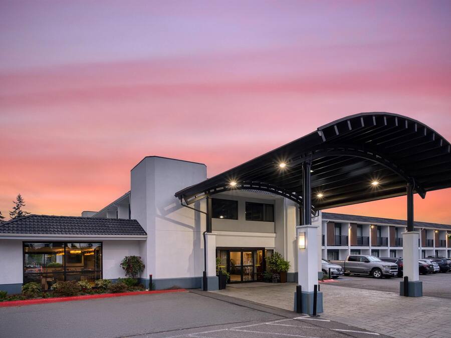 A hotel entrance with a covered waiting area, a parking lot, and a building with multiple windows.