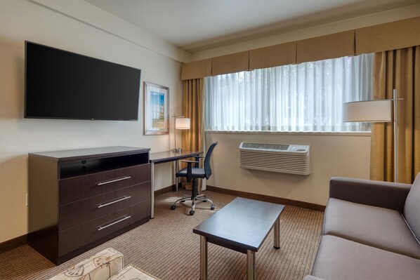 Habitación de hotel con televisión de pantalla plana, escritorio con silla, sofá, lámpara de pie y ventana con cortinas.