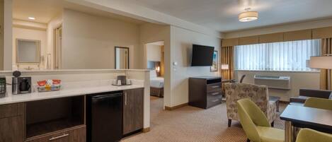 A hotel room with a kitchenette, a flat-screen TV, a dining area with chairs, and a large window.