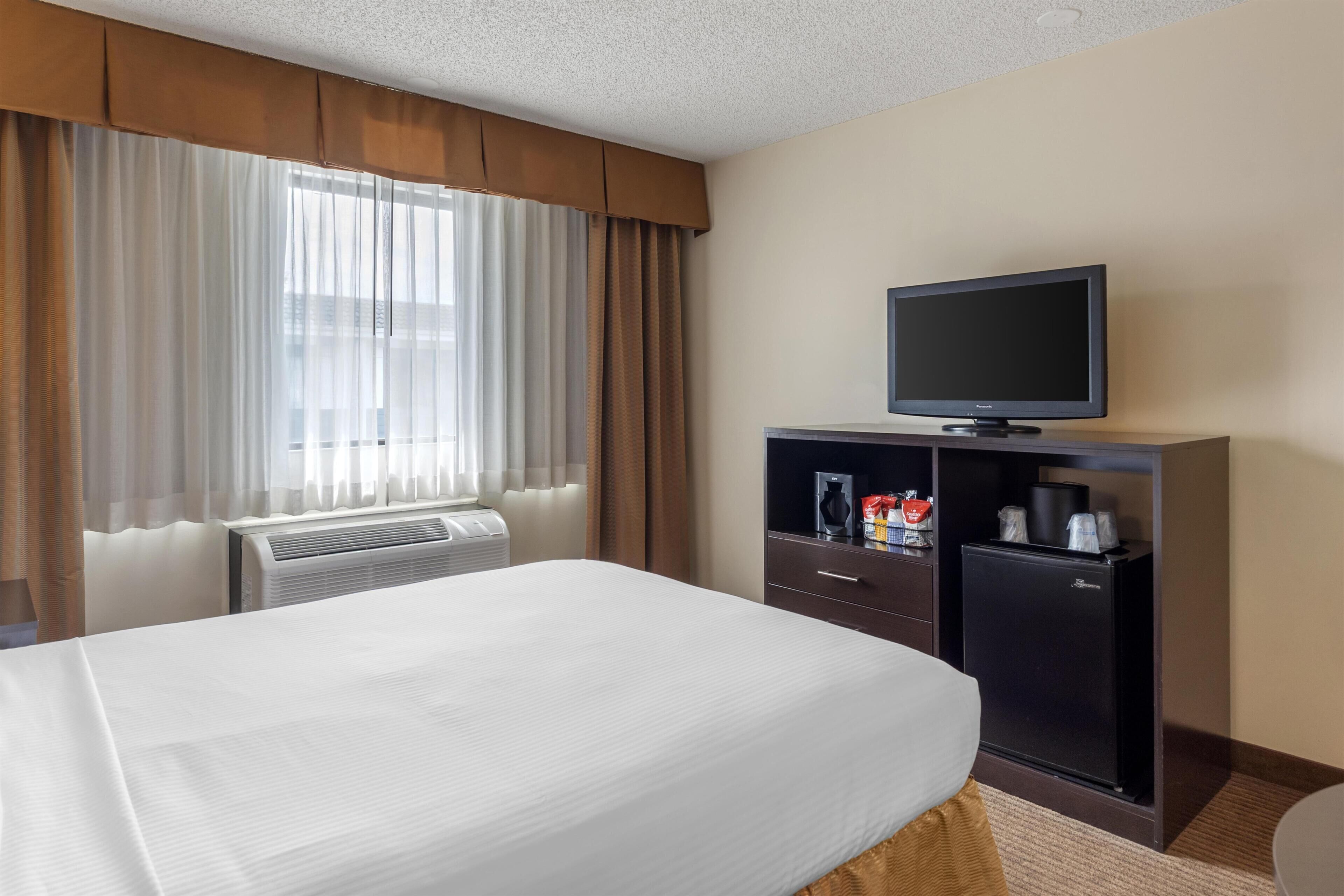 A hotel room with a bed, a television, a minibar, and a window with curtains.