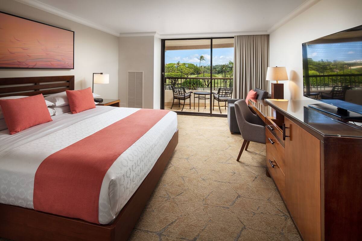 A hotel room with a large bed, a desk with a TV, a balcony with a view of greenery, and a chair.