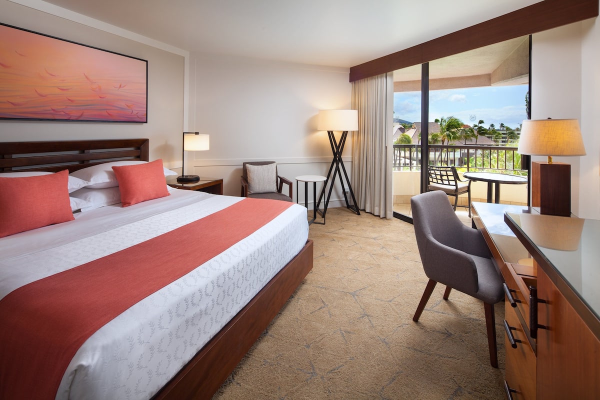 A hotel room with a large bed, a desk, a chair, a table, and a balcony with a view of palm trees.