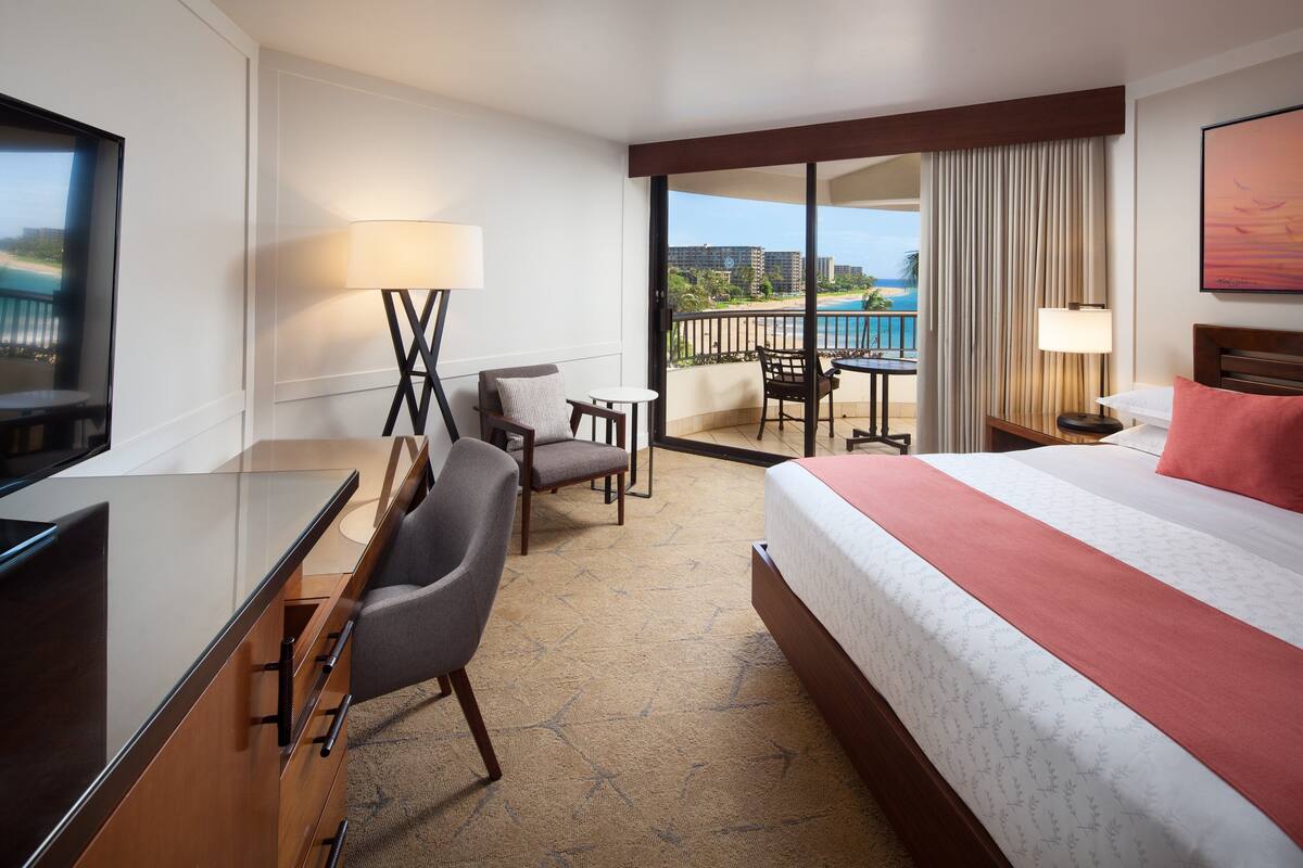 A hotel room with a bed, a desk, a chair, a small table, a TV, and a balcony with a view of the beach and buildings.