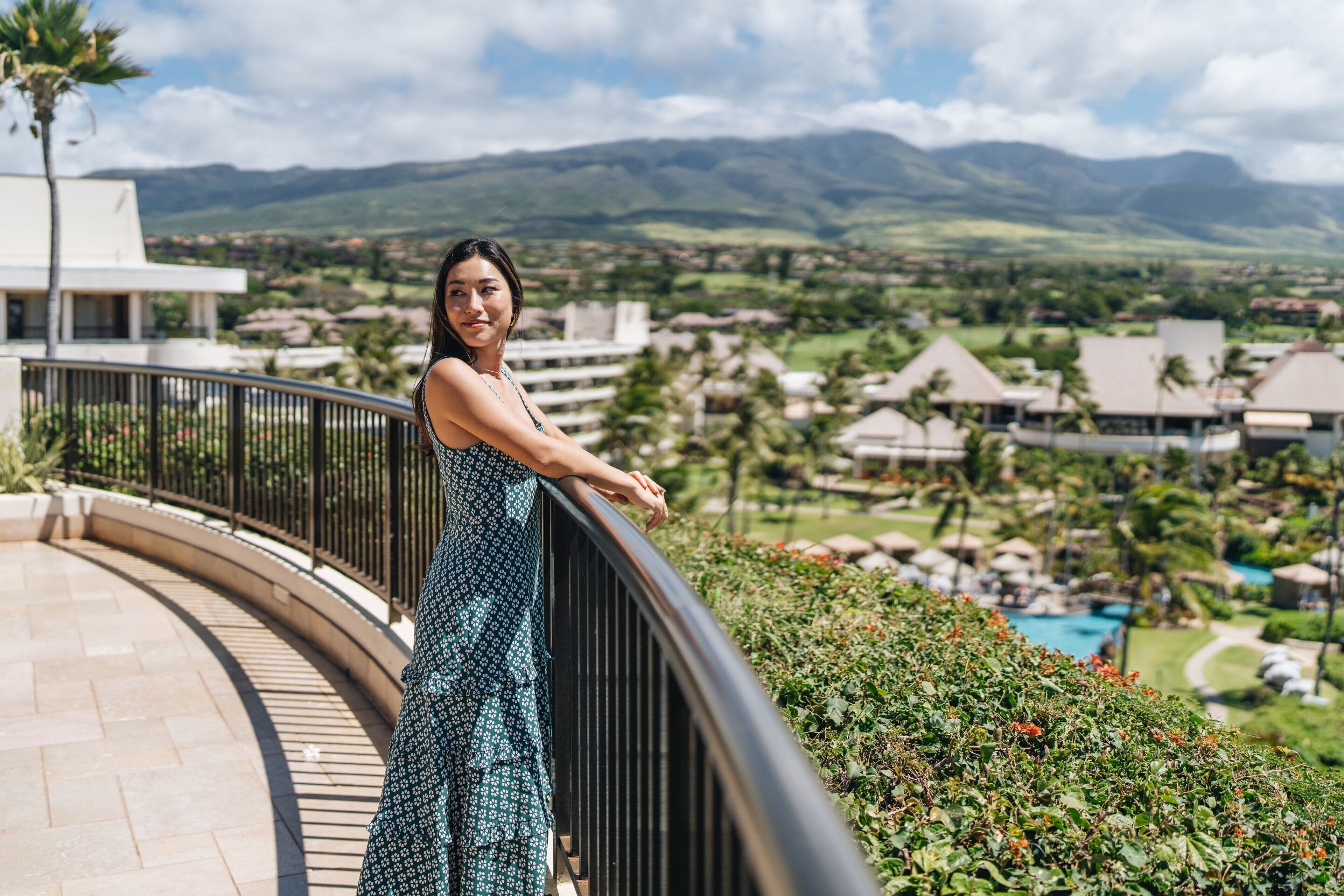 A person standing on a balcony with a view of a resort area, including buildings and a mountain range.