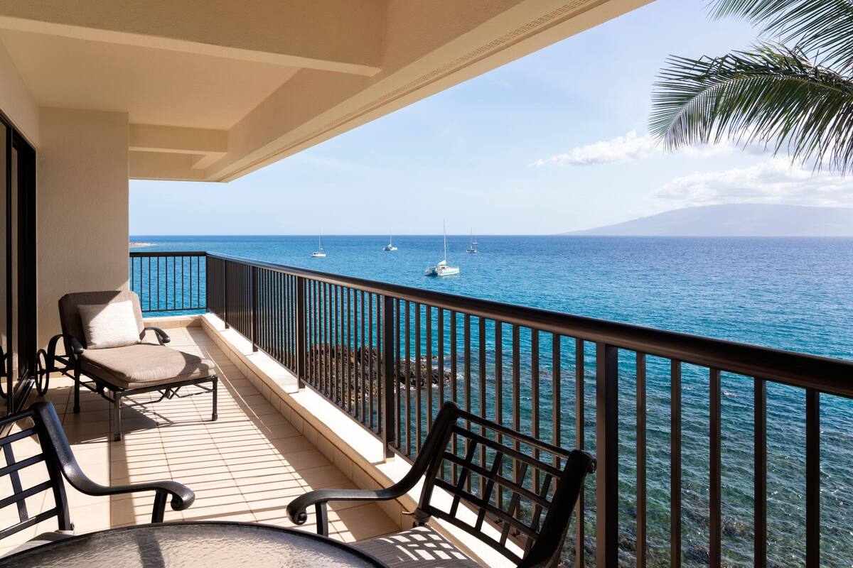 A balcony with two chairs and a table, overlooking a clear blue sea with several sailboats.