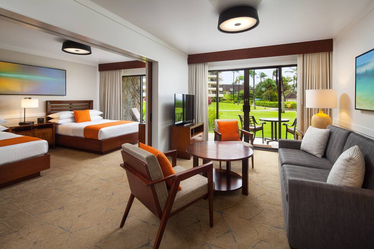 A hotel room with two beds, a sofa, a chair, a TV, and a balcony with a view of the garden.