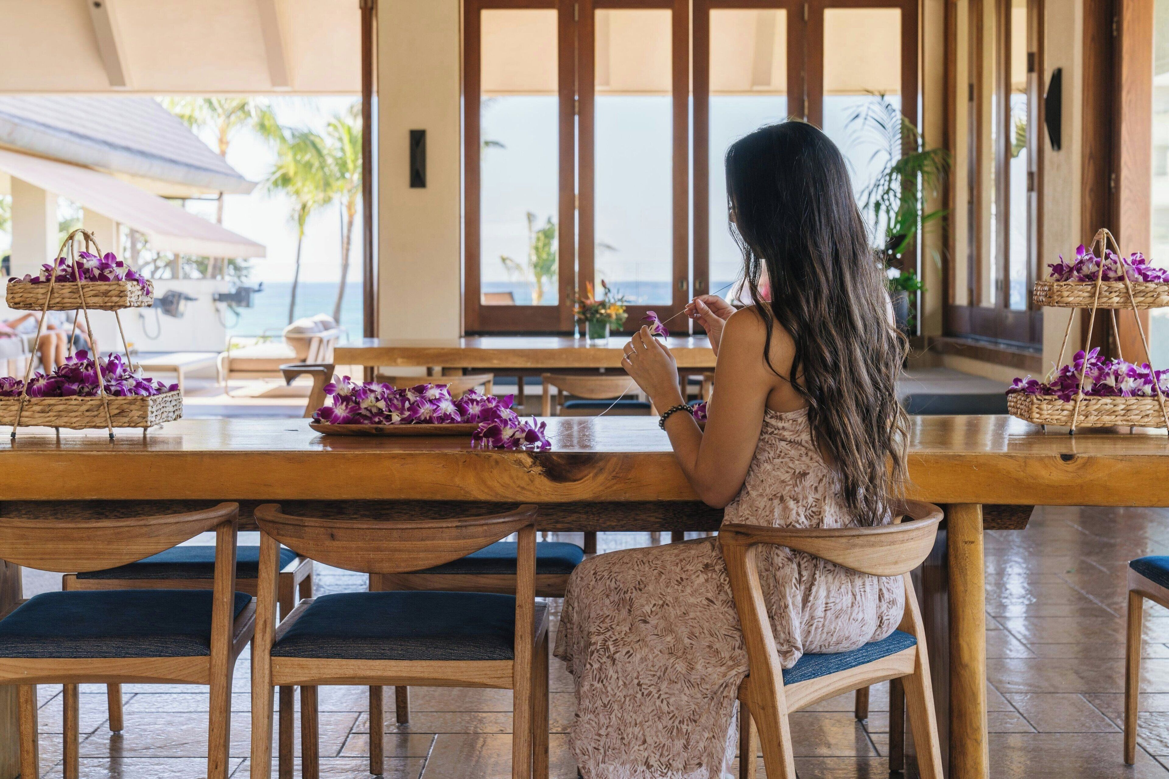 A woman sitting at a wooden table with purple flowers, overlooking a beach.