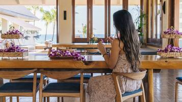 A woman sitting at a wooden table with purple flowers, overlooking a beach.