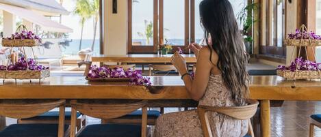 A woman sitting at a wooden table with purple flowers, overlooking a beach.